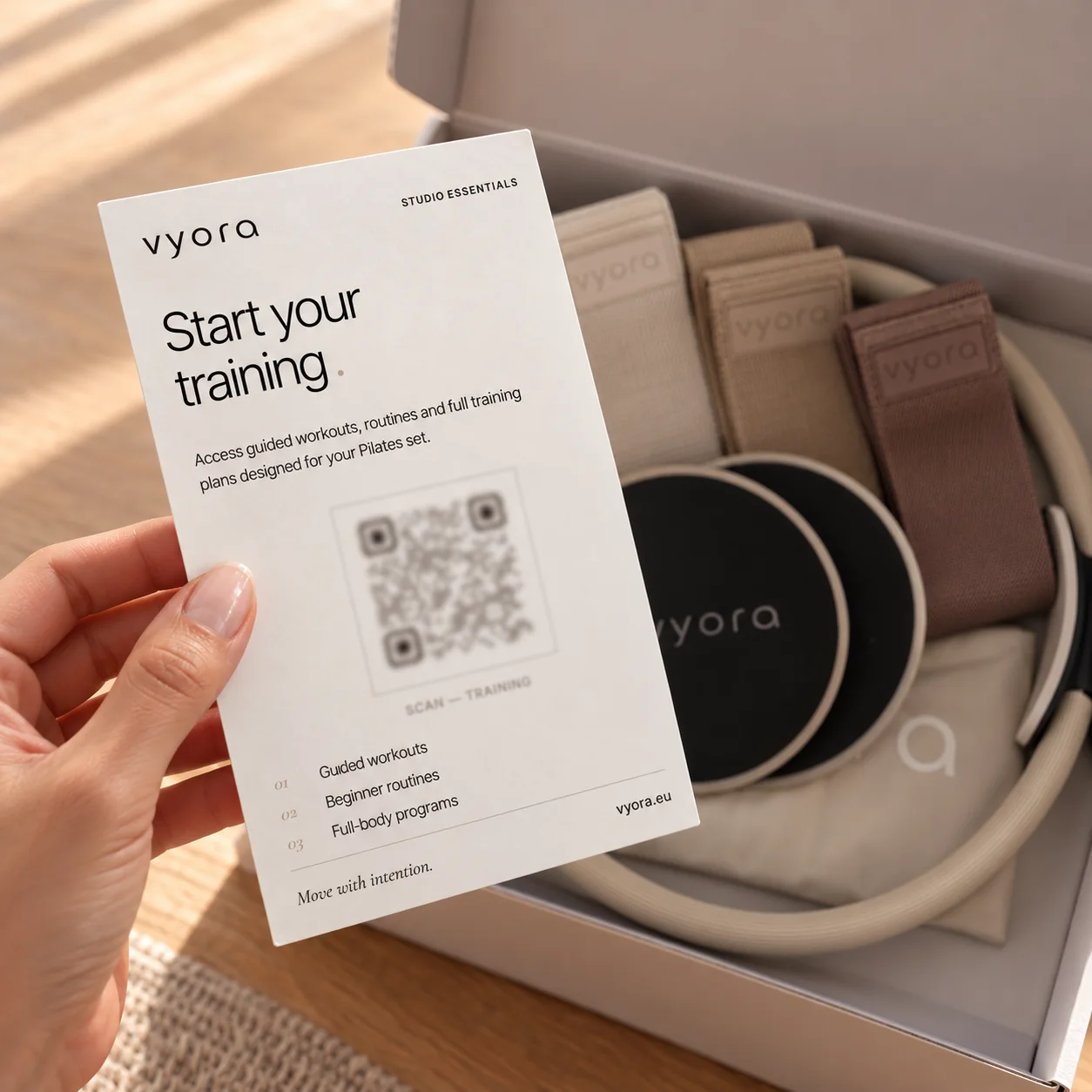 Eine Hand hält die vyora QR-Karte „Start your training“ über die geöffnete Verpackung mit Stoffbändern, Slidern und Ring